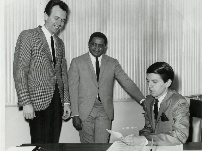 Eddie Ray standing between Bob Summers and Mike Curb, who sits at a desk, at MGM Records